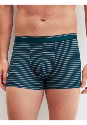 Männlicher Oberkörper, bekleidet mit dunkelgrünen Boxershorts mit dünnen rosa Horizontalstreifen, stehend mit entspannt an den Seiten hängenden Armen.