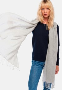 Femme blonde portant un pull bleu marine et un jean bleu tient une grande écharpe grise clair drapée sur ses épaules et étendue sur le côté.