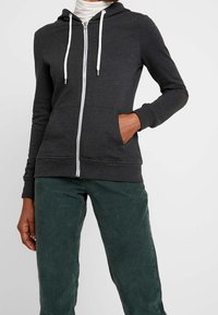 Osoba oblečená v tmavě šedé mikině na zip s bílými šňůrkami a zelených manšestrových kalhotách, s rukou v kapse, na jednobarevném pozadí.