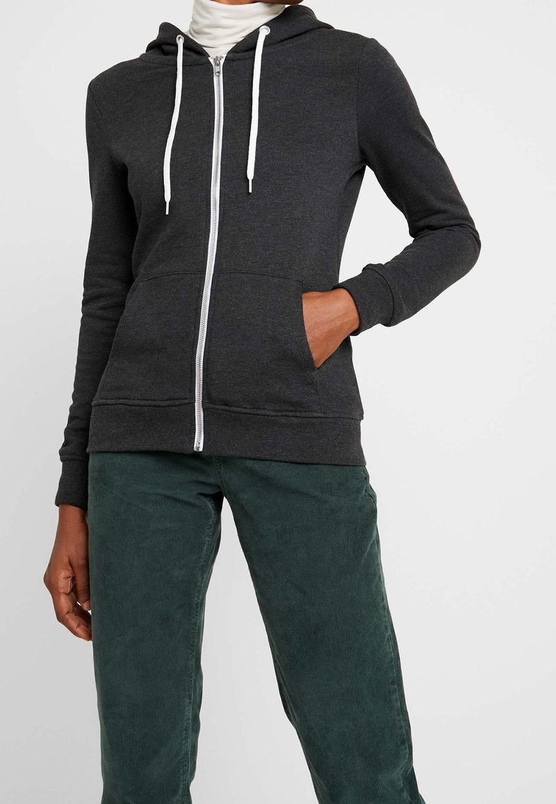 Osoba oblečená v tmavě šedé mikině na zip s bílými šňůrkami a zelených manšestrových kalhotách, s rukou v kapse, na jednobarevném pozadí.