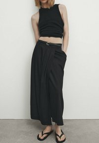Haut sans manches en tricot noir accompagné d'un pantalon noir taille haute à jambes larges avec des plis et une ceinture étroite. Porté avec des tongs noires.