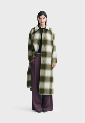 Femme portant un long manteau à carreaux verts et blancs par-dessus un haut bleu marine et un pantalon marron à larges jambes, debout devant un fond uni.