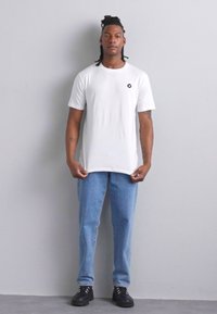 Vit bomulls t-shirt med korta ärmar och rund hals, med en liten svart cirkulär logotyp. Matchas med ljusblå denimjeans och svarta skor.