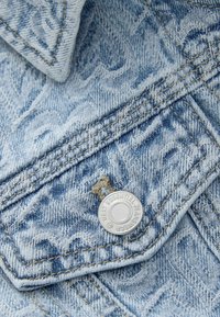 Helehallikas mustriga teksari kangas, topeltõmblusega õmblus ja hõbedane nupp, millel on kiri "NEXT ESSENTIAL JEANSWEAR."