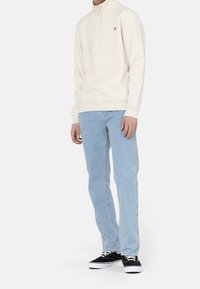 Sweatshirt a mezza zip crema con polsini a coste, abbinato a jeans azzurri. Il modello indossa scarpe nere. Design minimalista, senza motivi prominenti.