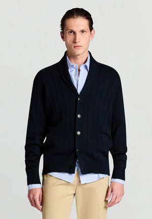 Marineblauer, gerippter Cardigan aus Strickstoff mit Schalkragen und Knopfverschluss, getragen über einem hellblau gestreiften Hemd.