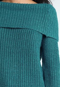 Pull en maille épaisse couleur bleu sarcelle avec un large décolleté largué sur les épaules, texture côtelée, et manches longues. Le tissu semble doux et confortable.