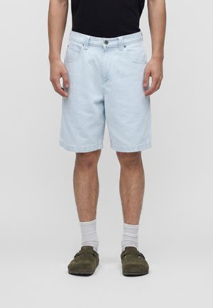 Mand iført lyseblå knælange cowboyshorts, hvide crew-sokker, olivengrønne slip-on sko og en sort t-shirt står foran en ensfarvet baggrund.