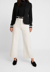 Blazer nero con polsini a righe, dolcevita nero, pantaloni beige a gamba larga e mocassini neri, che mostrano un aspetto moderno e sartoriale.