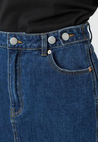 Sininen farkkuhame, jossa on korkea vyötärö, kaksinkertaiset hopeiset napit, oranssit ompeleet ja pieni etutasku.