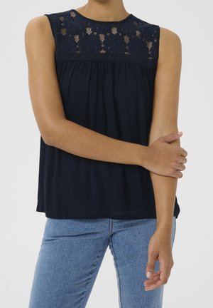 Femme portant un haut bleu marine sans manches avec un détail en dentelle au niveau de la poitrine et un jean bleu clair, tenant son bras gauche avec sa main droite.