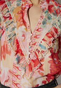 Blouse fleurie avec col froncé et finition en dentelle. Présente des motifs vibrants rouges, roses et aqua. Tissu léger avec des boutons près de l'encolure.