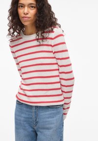 Πλεκτό πουλόβερ με ρίγες κόκκινες και λευκές, στρογγυλή λαιμόκοψη και μακριά μανίκια, συνδυασμένο με μπλε τζιν τζιν. Χαλαρή εφαρμογή και υφασμάτινη υφή.