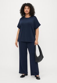 Ναυτικό μπλε χαλαρό μπλουζάκι με διπλωμένα κοντά μανίκια, συνδυασμένο με ένα παντελόνι με φαρδιά εφαρμογή. Το μοντέλο κρατάει μια μαύρη τσάντα, φορώντας μαύρα παπούτσια.