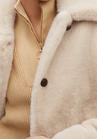 Cremefarvet, furry stofjakke med notched krave, prydet med brune knapper og en blød tekstur, lagdelt over en beige strikket top.