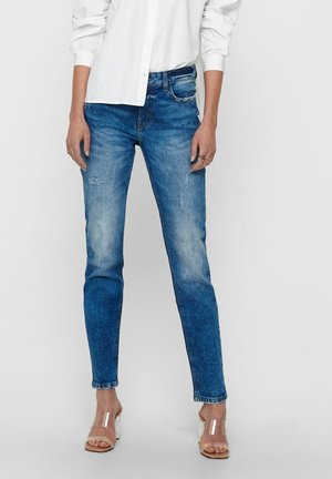 Jeans ajustados de denim azul, con un lavado desgastado y detalles rasgados. Combinados con sandalias de tacón transparente y una camisa blanca de botones.