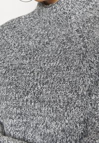 Grå stickad tröja med hög krage, texturerat mönster och varierande nyanser av mörk och ljusgrå garn. Mjuk och mysig känsla.