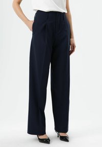 Pantalon large marine taille haute, devant plissé, poches latérales et fini en tissu lisse, assorti à des talons noirs pointus.