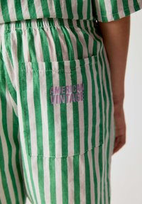Pantalon à rayures verticales vertes et blanches avec une poche arrière portant un texte violet indiquant American Vintage sur le côté droit.