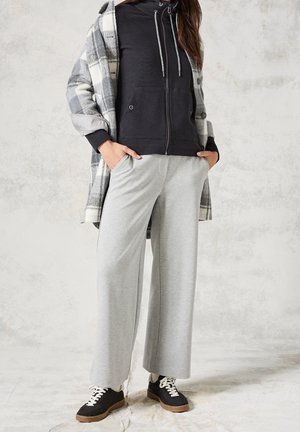 Woman wearing gray wide-leg pants, black zip-up hoodie, gray plaid coat, and black sneakers with white laces, standing with hands in pockets.