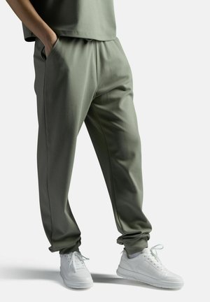 Ihmisen alaosa, jolla oli oliivinvihreät collegehousut ja valkoiset lenkkarit, kädet taskuissa yksiväristä taustaa vasten.