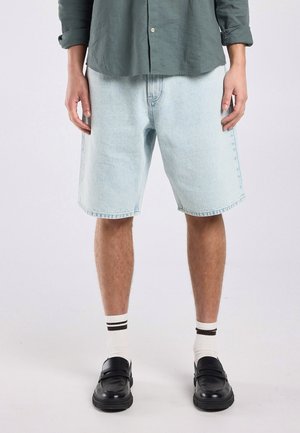 Man die lichtblauwe denimshorts, witte sokken met zwarte strepen, zwarte loafers en een groene lange mouw shirt draagt, staand tegen een effen achtergrond.
