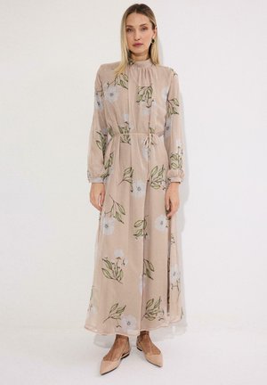 Kobieta ubrana w długą beżową sukienkę w kwiaty z długimi rękawami i wiązanym w pasie paskiem, w połączeniu z beżowymi, spiczastymi butami, stojąca na jednolitym tle.