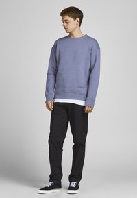 Sudadera morada con cuello redondo y mangas largas, superpuesta sobre una camisa blanca. Combinada con pantalones negros y zapatillas negras.