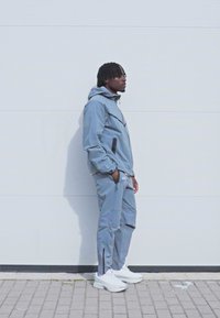 Pelēks sporta žakets ar kapuci un rāvējslēdzēja kabatām, atbilstošas bikses ar sānu rāvējslēdzējiem un baltas sporta apavus. Gluda tekstūra, minimālists dizains.