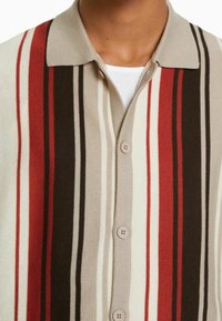 Gebreide shirt met een button-down kraag, met verticale strepen in rood, beige, bruin en crème. Zachte textuur met vier zichtbare knopen.