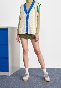 Stickad väst i krämfärg med blåa detaljer och knappar, kombinerad med en grön minikjol. Grå sneakers med ljusa sulor och vita strumpor. Blå byrå.