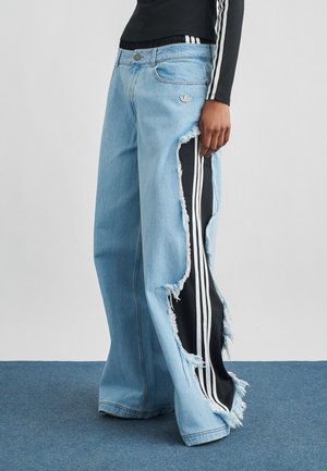 Személy világoskék, széles szárú farmernadrágot visel, nagy oldalsó kivágásokkal, amelyek alatt fekete anyag látszik fehér csíkokkal.