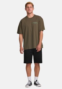 Olive green cotton t-shirt with short sleeves and a crew neck, featuring a small brand logo in green on the chest. Paired with black shorts.