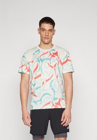 Sports t-skjorte i lys beige med et virvlende mønster i rød og teal. Har korte ermer og rund hals, laget av lett stoff.
