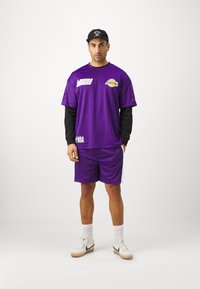 Camiseta deportiva morada con textura de malla, con logotipos del equipo. Combinada con pantalones cortos morados y una gorra negra, complementada con zapatillas blancas.