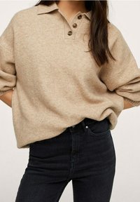 Suéter de color beige, de punto, con cuello con solapa, tres botones y mangas cortas ligeramente dobladas, combinado con jeans oscuros.