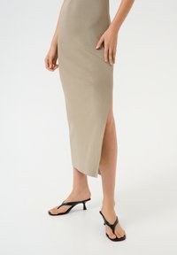 Beige, knielanges Strickkleid mit seitlichem Schlitz, kombiniert mit schwarzen, trägerlosen Sandalen mit niedrigem Absatz. Glatte Textur und figurbetontes Design.