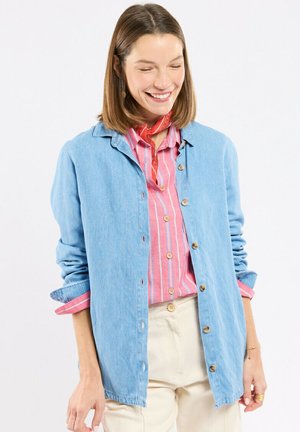 Femme souriante aux cheveux bruns mi-longs porte une veste bleue boutonnée sur une chemise rayée rose et un pantalon crème.