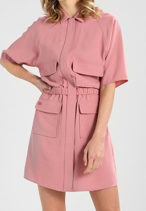 Woman wearing a short-sleeve pink utility dress with four large flap pockets and an elastic waistband against a plain background.