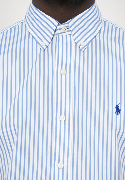 Svijetloplava i bijela prugasta košulja s kopčanjem na gumbe, šiljastim ovratnikom, bijelim gumbima i malim plavim logotipom na lijevoj strani.