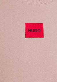 Tissu rose clair avec un écusson carré rouge cousu, affichant le mot HUGO en lettres majuscules noires.