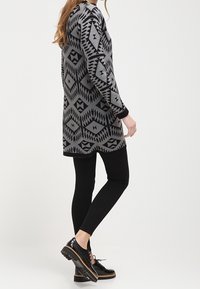 Tunique grise à manches longues avec motifs géométriques noirs ; assortie à des leggings noirs et des chaussures noires brillantes à semelle texturée.