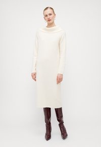 Langermet, kremfarget strikket kjole med cowl-neck. Glatt tekstur, rett passform, knelang, kombinert med blanke burgunder knelengde støvler.
