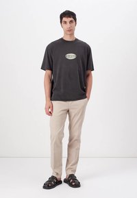 Mørk grå t-skjorte med en sentral grønn og hvit "GUESS" logo, kombinert med lyse beige bukser og svarte sandaler med flere stropper.