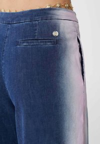 Pantaloni in denim con un design a gradiente che va dal blu scuro al rosa chiaro, dotati di una tasca posteriore e dettagli in metallo argentato. Texture liscia.