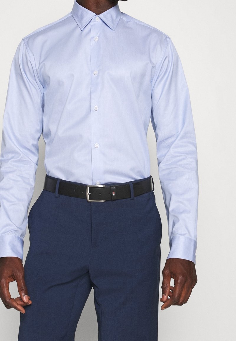 Světlemodrá košile na knoflíky s špičatým límcem, dlouhými rukávy a výraznou strukturou. V kombinaci s tmavě modrými kalhotami a černým opaskem.