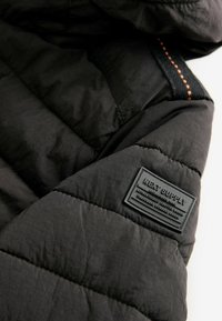 Giacca piumino nera con una texture trapuntata. Presenta un filo di cucitura arancione e un'etichetta in gomma sul maniche sinistro.