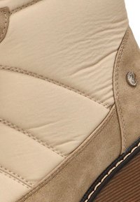 Botín beige hasta el tobillo, hecho de cuero texturizado, con costuras decorativas y un pequeño logotipo plateado. La suela es de color marrón oscuro con surcos.