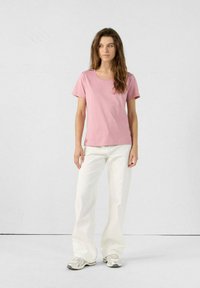Camiseta de algodón rosa claro con mangas cortas y cuello redondo. Combinada con pantalones blancos de corte holgado y zapatillas deportivas plateadas. Fondo neutro.