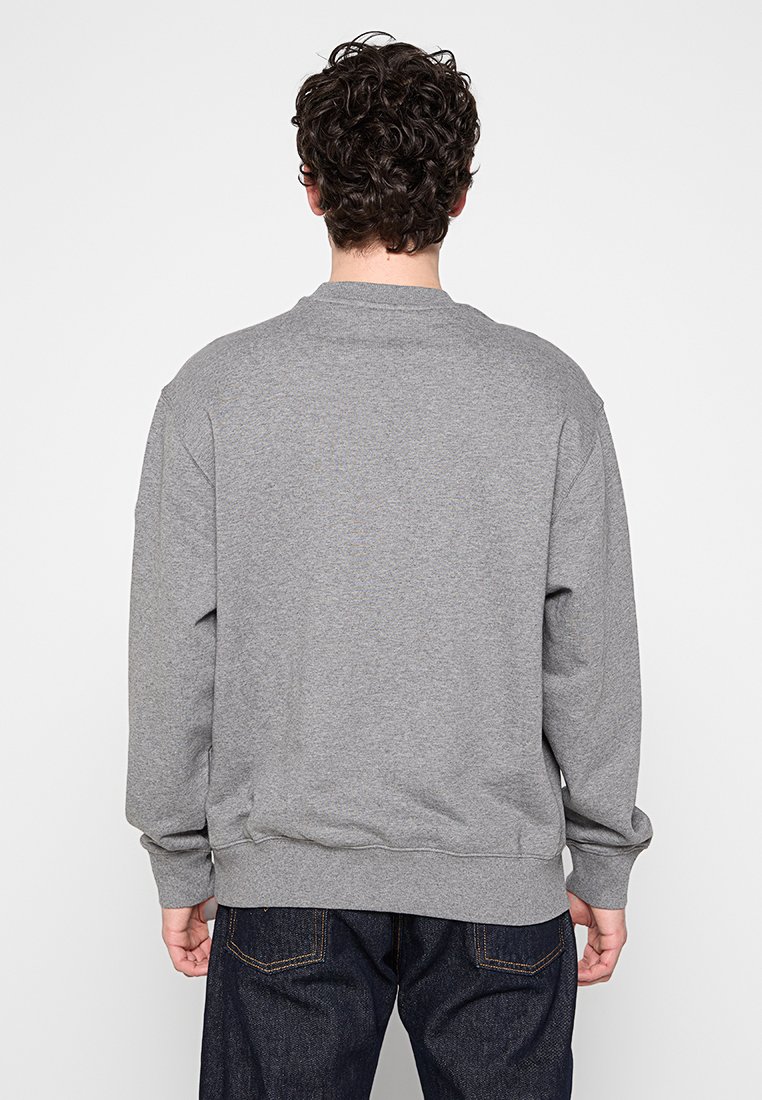 Hombre con cabello rizado que lleva un sudadera gris lisa y jeans azul oscuro, visto desde atrás sobre un fondo blanco.